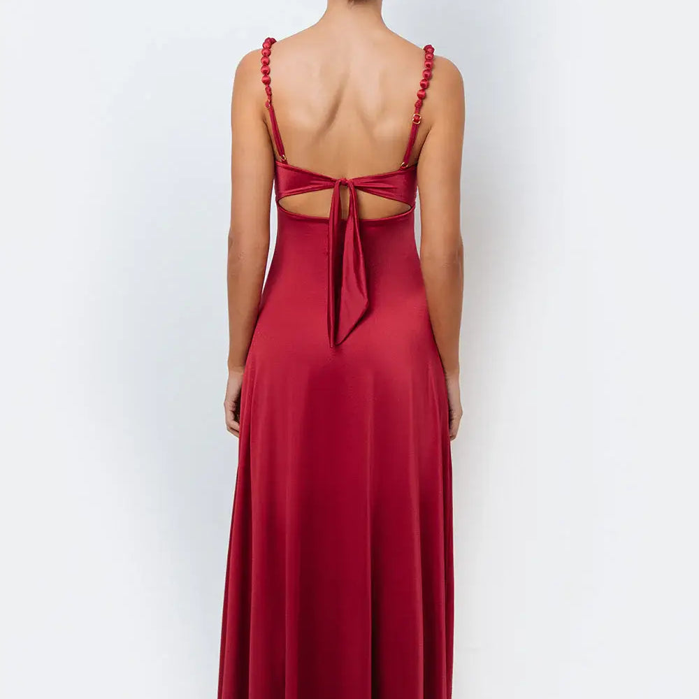 A woman modeling a deep crimson, flowy maxi dress with a sweetheart knotted bodice, center cutout, pearl-accented straps, and a back tie.