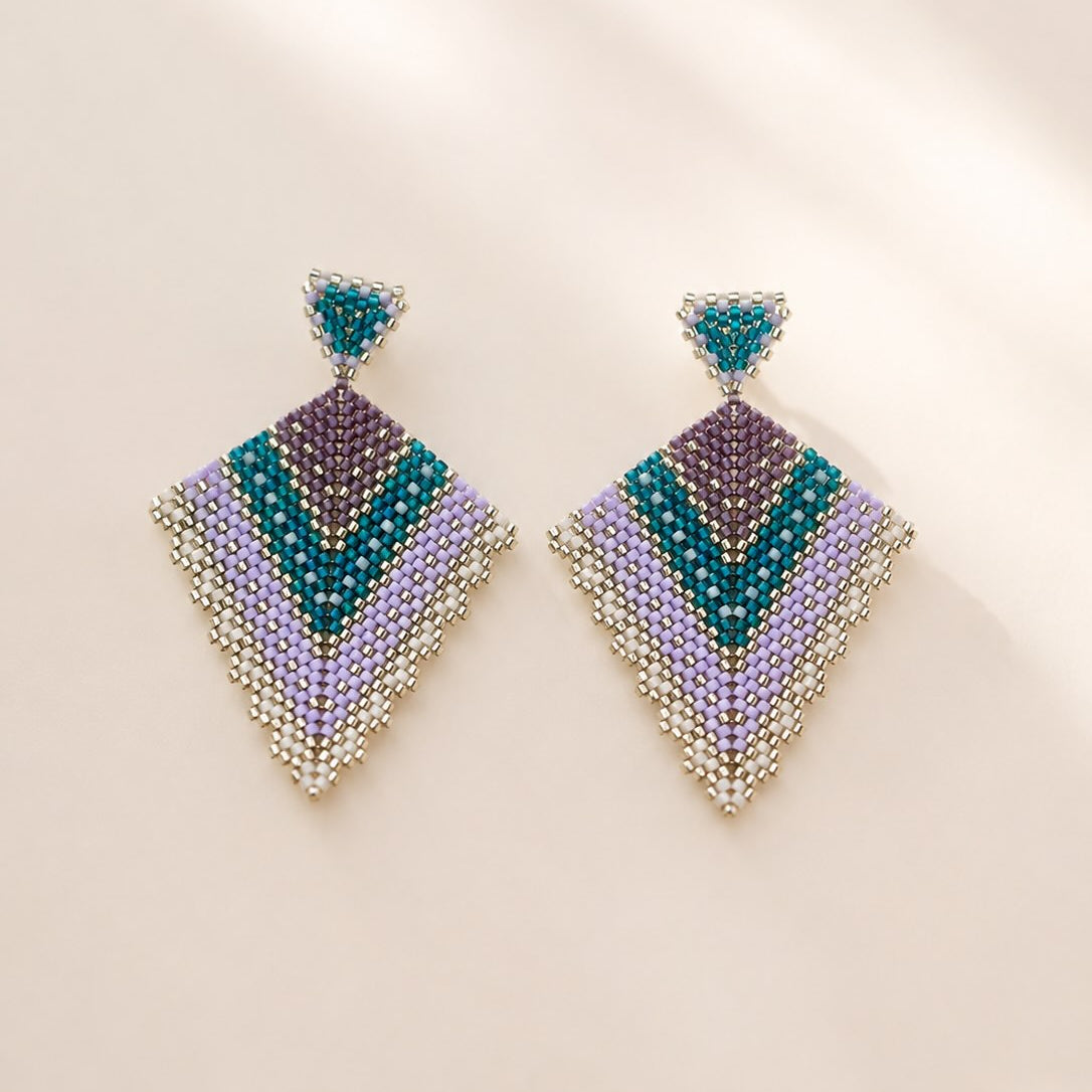 A pair of geometric beaded drop earrings with a chevron pattern in teal, purple, lavender, and silver, displayed on a warm beige background with soft shadows.