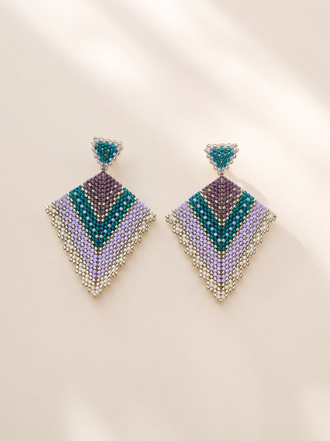 A pair of geometric beaded drop earrings with a chevron pattern in teal, purple, lavender, and silver, displayed on a warm beige background with soft shadows.