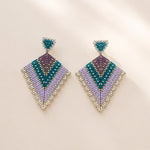 A pair of geometric beaded drop earrings with a chevron pattern in teal, purple, lavender, and silver, displayed on a warm beige background with soft shadows.