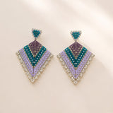 A pair of geometric beaded drop earrings with a chevron pattern in teal, purple, lavender, and silver, displayed on a warm beige background with soft shadows.