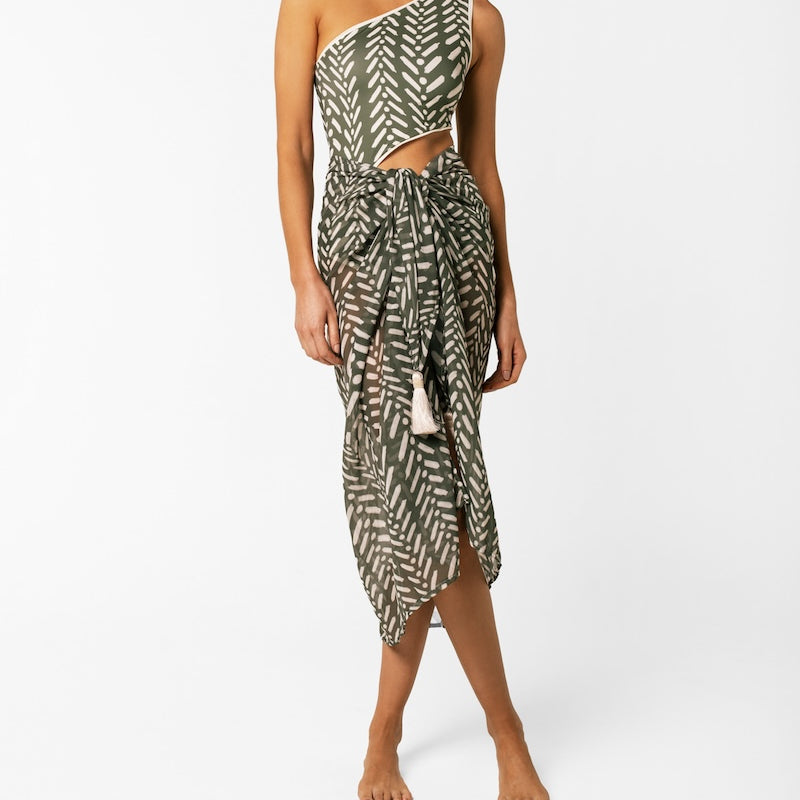 Woman wearing a green and white patterned one-shoulder one-piece bikini with a matching shawl on a white background. 