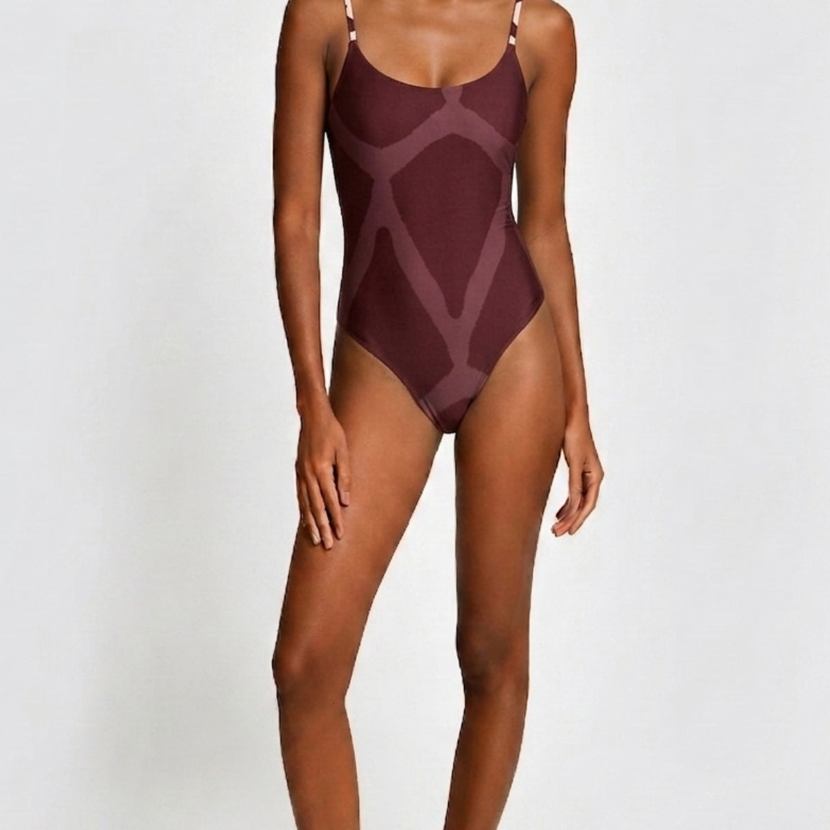 A woman modeling a one-piece swimsuit with a geometric pattern in deep burgundy and muted mauve tones, set against a plain light gray studio background.