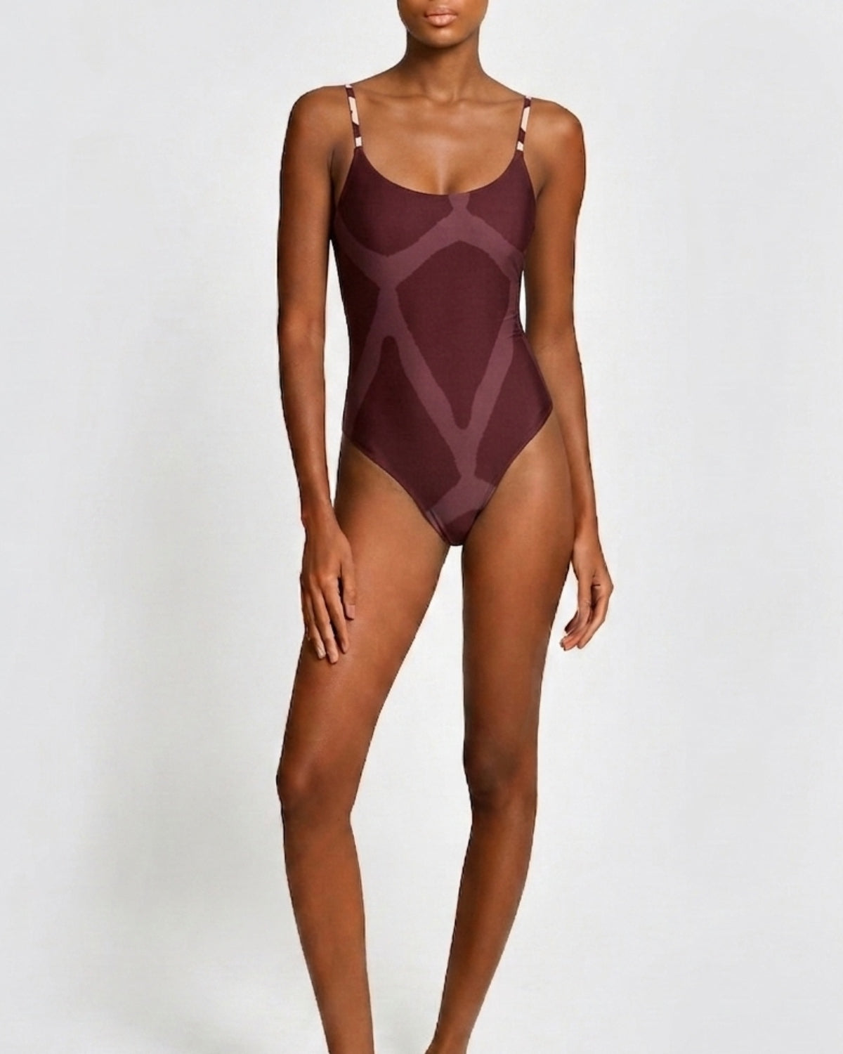 A woman modeling a one-piece swimsuit with a geometric pattern in deep burgundy and muted mauve tones, set against a plain light gray studio background.