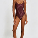A woman modeling a one-piece swimsuit with a geometric pattern in deep burgundy and muted mauve tones, set against a plain light gray studio background.