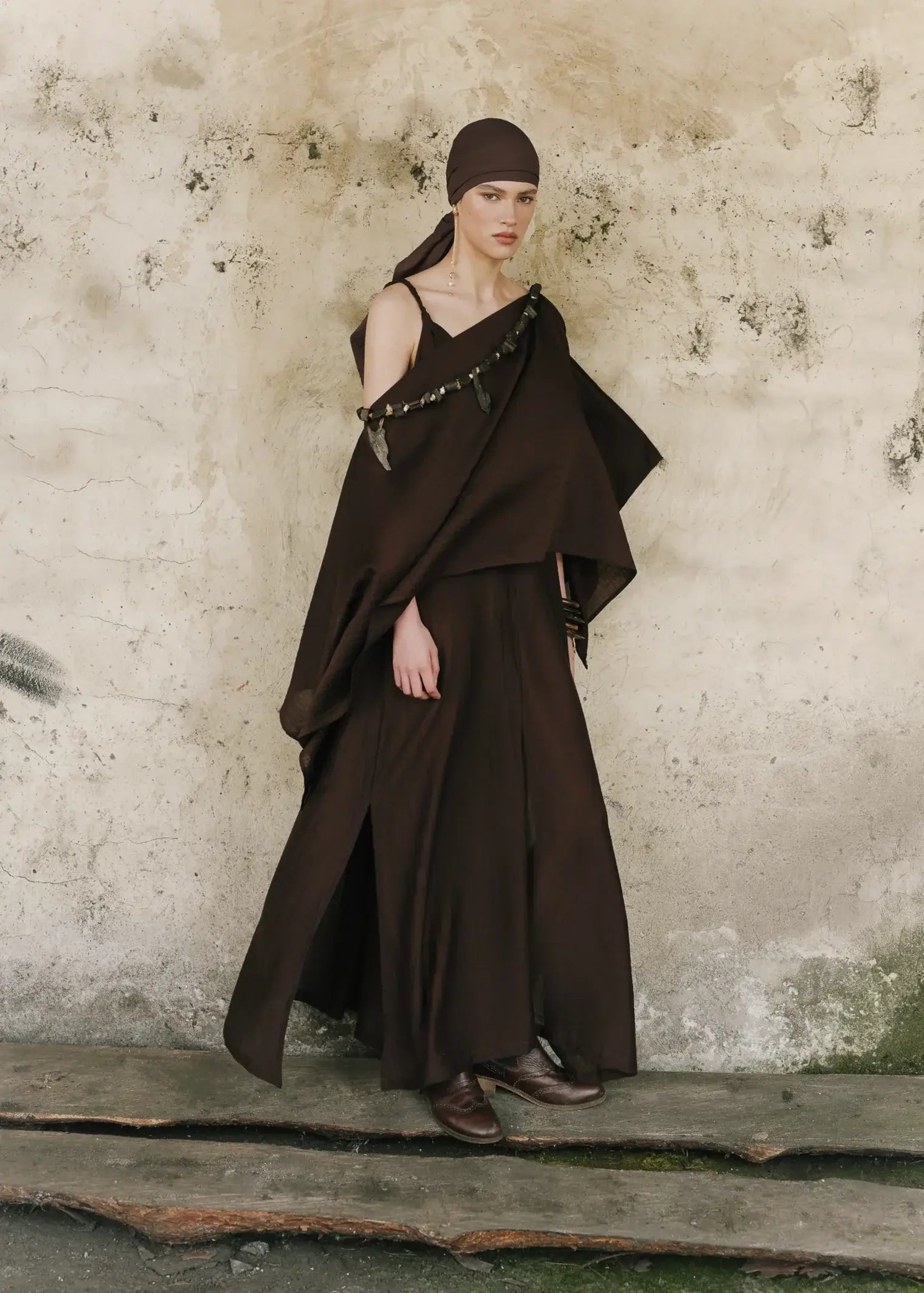Woman wearing a dark brown dress and headscarf against a textured wall.