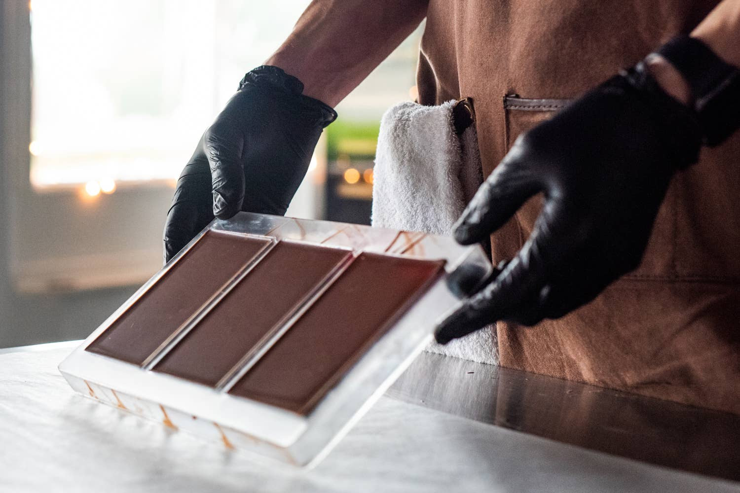 Person wearing black gloves and a brown apron arranging chocolate bars on a white surface.