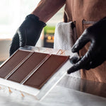Person wearing black gloves and a brown apron arranging chocolate bars on a white surface.
