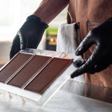 Person wearing black gloves and a brown apron arranging chocolate bars on a white surface.