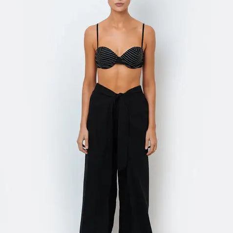 A woman standing against a white background wearing black resortwear pants with a tie-front detail and a matching black bikini top.