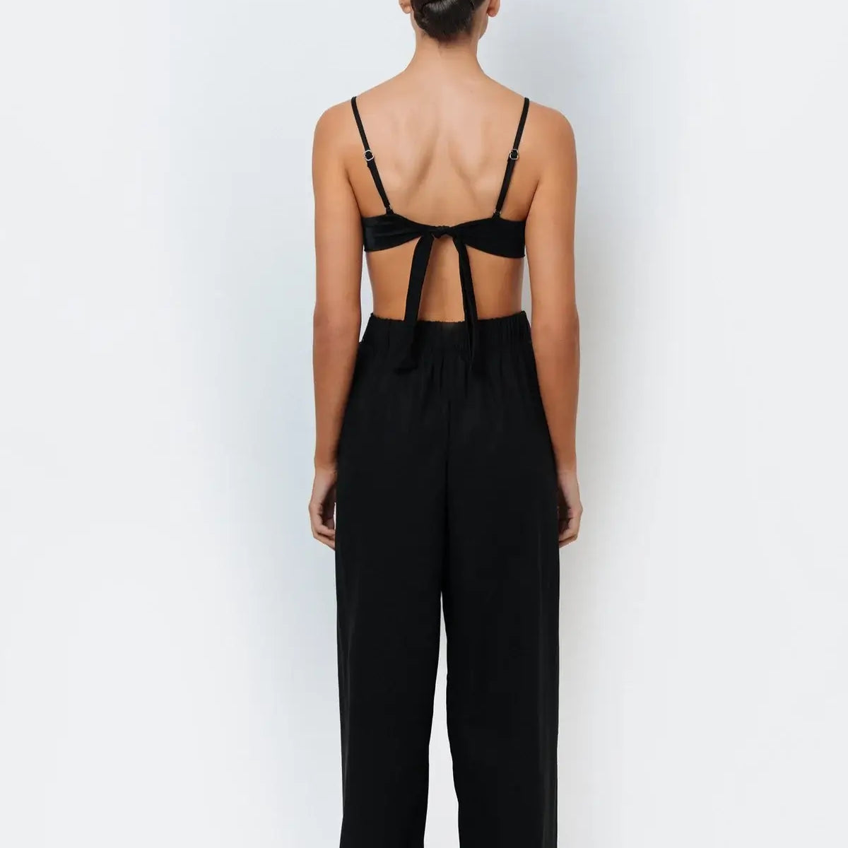 A woman standing against a white background wearing black resortwear pants with a tie-front detail and a matching black bikini top.