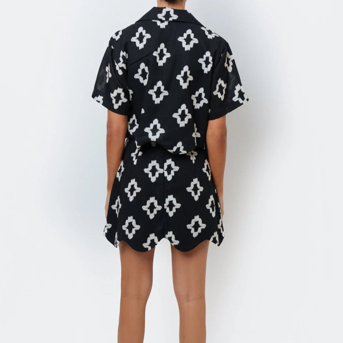 A woman standing against a white background wearing a black and white geometric patterned button down shirt with scalloped hem detail and a matching skirt.