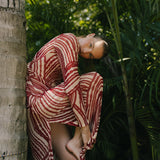 Woman in a red and white patterned dress leaning against a tree in a tropical setting.