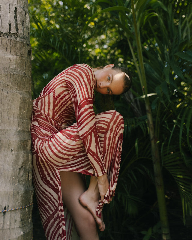 Woman in a red and white patterned dress leaning against a tree in a tropical setting.