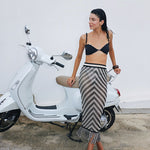 Woman in a black bikini top and patterned skirt standing next to a white scooter against a white wall.