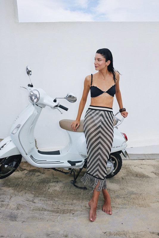 Woman in a black bikini top and patterned skirt standing next to a white scooter against a white wall.