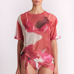 A model wearing a sheer pink and white floral print swim cover-up top with a side drawstring, paired with matching bikini bottoms, against a plain light background.