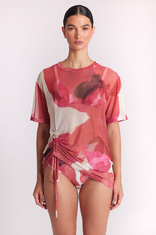 A model wearing a sheer pink and white floral print swim cover-up top with a side drawstring, paired with matching bikini bottoms, against a plain light background.