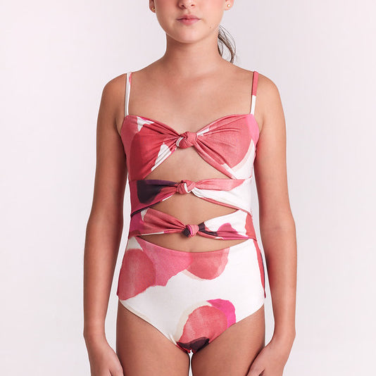 A girl wearing a one-piece swimsuit with a pink and white abstract floral pattern, featuring a bow detail on the bust and a lace-up front design, set against a plain light background.