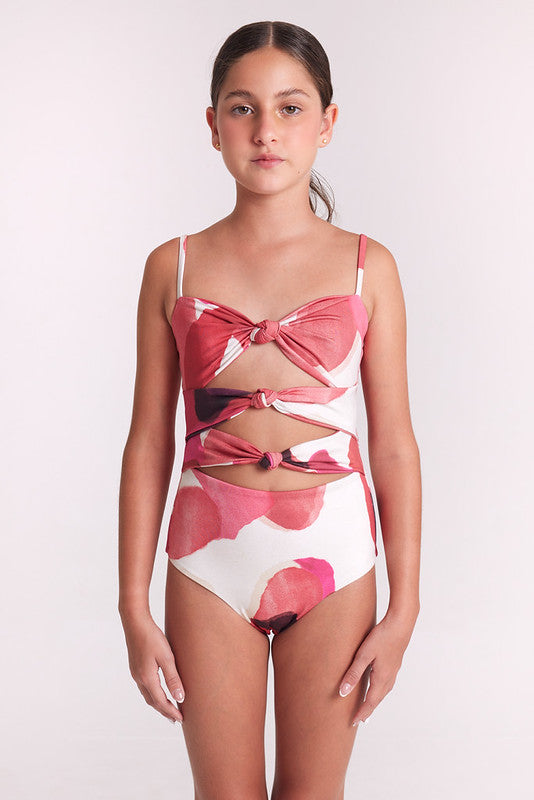 A girl wearing a one-piece swimsuit with a pink and white abstract floral pattern, featuring a bow detail on the bust and a lace-up front design, set against a plain light background.