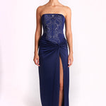 A model stands against a plain white studio background wearing a strapless navy maxi dress with a fitted bodice, light metallic embroidery, and a gathered skirt with a high front slit.