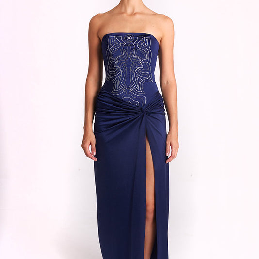 A model stands against a plain white studio background wearing a strapless navy maxi dress with a fitted bodice, light metallic embroidery, and a gathered skirt with a high front slit.