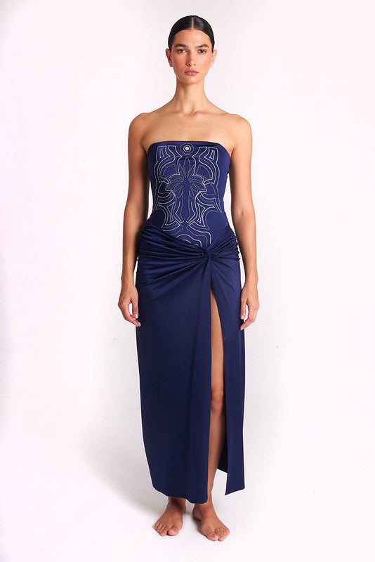 A model stands against a plain white studio background wearing a strapless navy maxi dress with a fitted bodice, light metallic embroidery, and a gathered skirt with a high front slit.