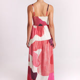 A woman viewed from behind wearing a sleeveless maxi dress with thin straps and an abstract floral pattern in pink, coral, red, and cream, photographed against a plain light background.
