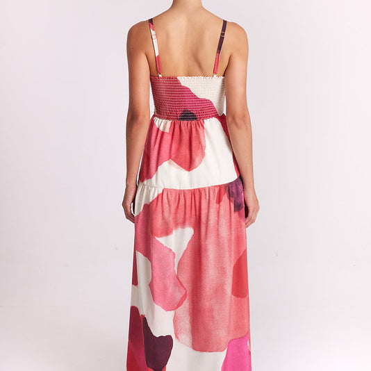 A woman viewed from behind wearing a sleeveless maxi dress with thin straps and an abstract floral pattern in pink, coral, red, and cream, photographed against a plain light background.