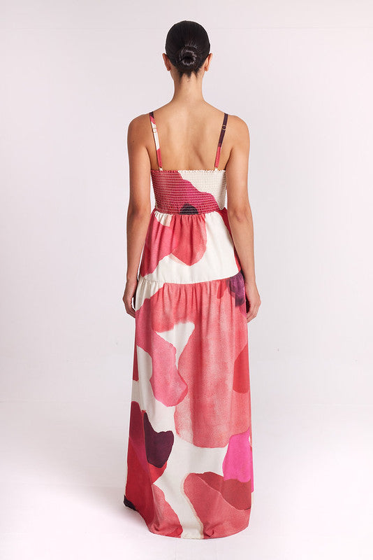 A woman viewed from behind wearing a sleeveless maxi dress with thin straps and an abstract floral pattern in pink, coral, red, and cream, photographed against a plain light background.