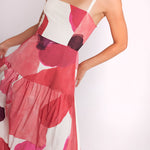 A woman wearing a sleeveless tiered maxi dress with a large abstract watercolor print in pink, red, and white, photographed against a plain white studio background.