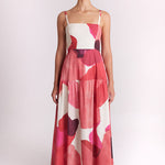 A woman modeling a sleeveless tiered maxi dress with a pink, red, and white abstract floral pattern against a plain light background.