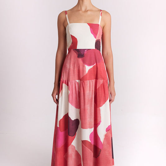 A woman modeling a sleeveless tiered maxi dress with a pink, red, and white abstract floral pattern against a plain light background.