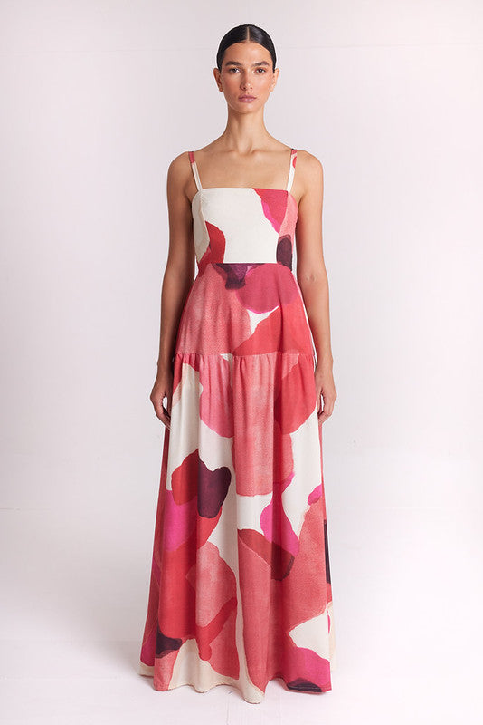 A woman modeling a sleeveless tiered maxi dress with a pink, red, and white abstract floral pattern against a plain light background.