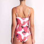 A woman viewed from behind wearing a one-piece swimsuit with a pink and red abstract floral pattern on a white background, set against a plain light gray studio backdrop.