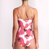 A woman viewed from behind wearing a one-piece swimsuit with a pink and red abstract floral pattern on a white background, set against a plain light gray studio backdrop.