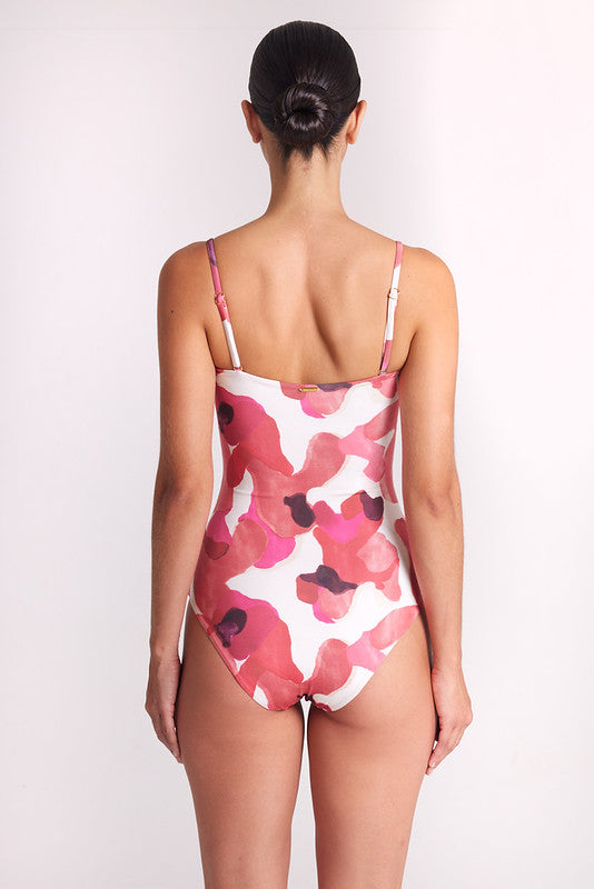 A woman viewed from behind wearing a one-piece swimsuit with a pink and red abstract floral pattern on a white background, set against a plain light gray studio backdrop.