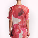 A woman viewed from behind wearing a sheer short-sleeve tunic top with a pink, red, and white abstract floral pattern, set against a plain white studio background.