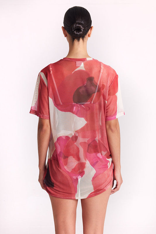 A woman viewed from behind wearing a sheer short-sleeve tunic top with a pink, red, and white abstract floral pattern, set against a plain white studio background.