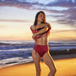 A woman posing on a sandy beach at sunset wearing a red two-piece bikini, with ocean waves and a colorful sky in the background.
