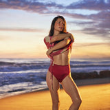 A woman posing on a sandy beach at sunset wearing a red two-piece bikini, with ocean waves and a colorful sky in the background.