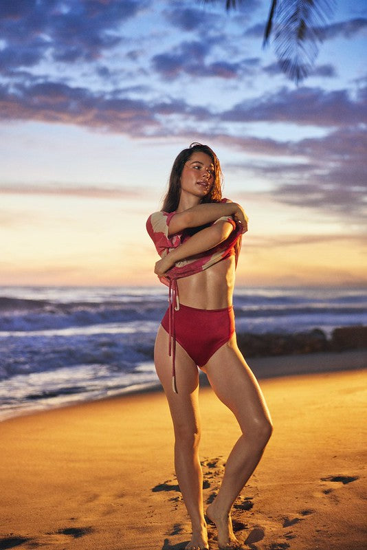 A woman posing on a sandy beach at sunset wearing a red two-piece bikini, with ocean waves and a colorful sky in the background.