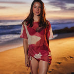 A woman stands on a sandy beach at sunset wearing a loose, short dress with a red, pink, and cream abstract floral pattern. The ocean and warm sky are visible in the background.