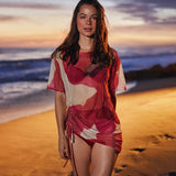 A woman stands on a sandy beach at sunset wearing a loose, short dress with a red, pink, and cream abstract floral pattern. The ocean and warm sky are visible in the background.