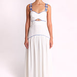 A model stands against a plain light background wearing a white sleeveless maxi dress with blue trim and blue rope details on the straps.