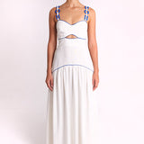 A model stands against a plain light background wearing a white sleeveless maxi dress with blue trim and blue rope details on the straps.