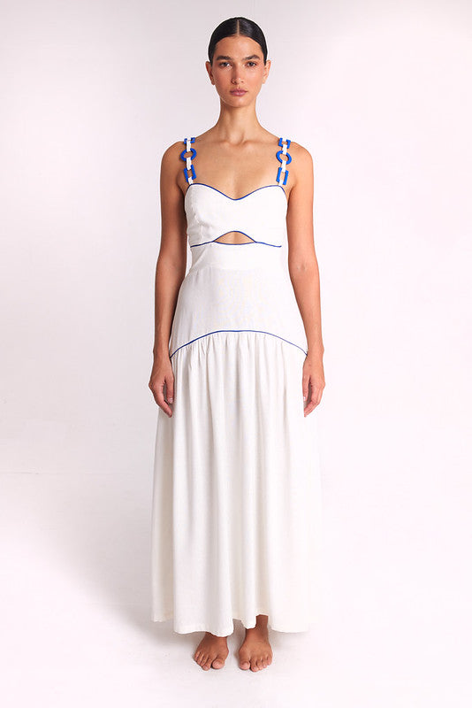 A model stands against a plain light background wearing a white sleeveless maxi dress with blue trim and blue rope details on the straps.
