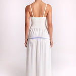 A woman viewed from the back wearing a white maxi dress with thin straps, a smocked bodice, and a thin blue horizontal stripe at the waist, set against a plain light background.