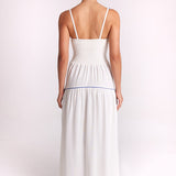A woman viewed from the back wearing a white maxi dress with thin straps, a smocked bodice, and a thin blue horizontal stripe at the waist, set against a plain light background.