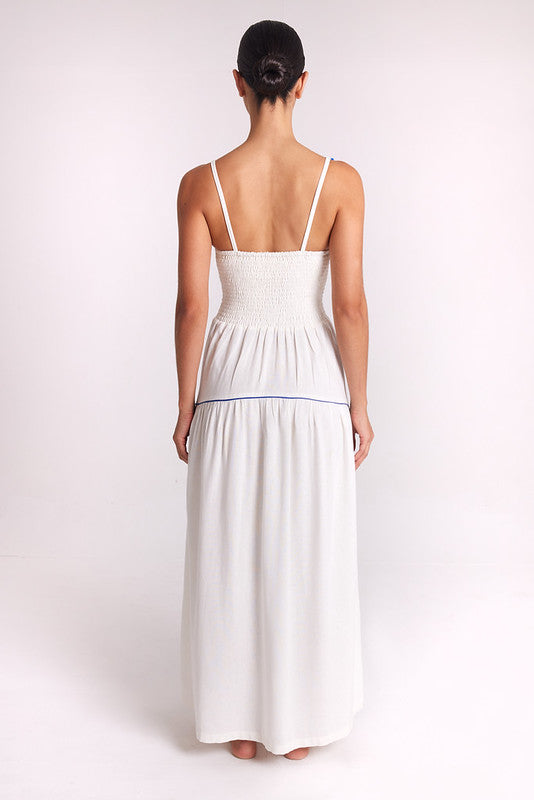 A woman viewed from the back wearing a white maxi dress with thin straps, a smocked bodice, and a thin blue horizontal stripe at the waist, set against a plain light background.